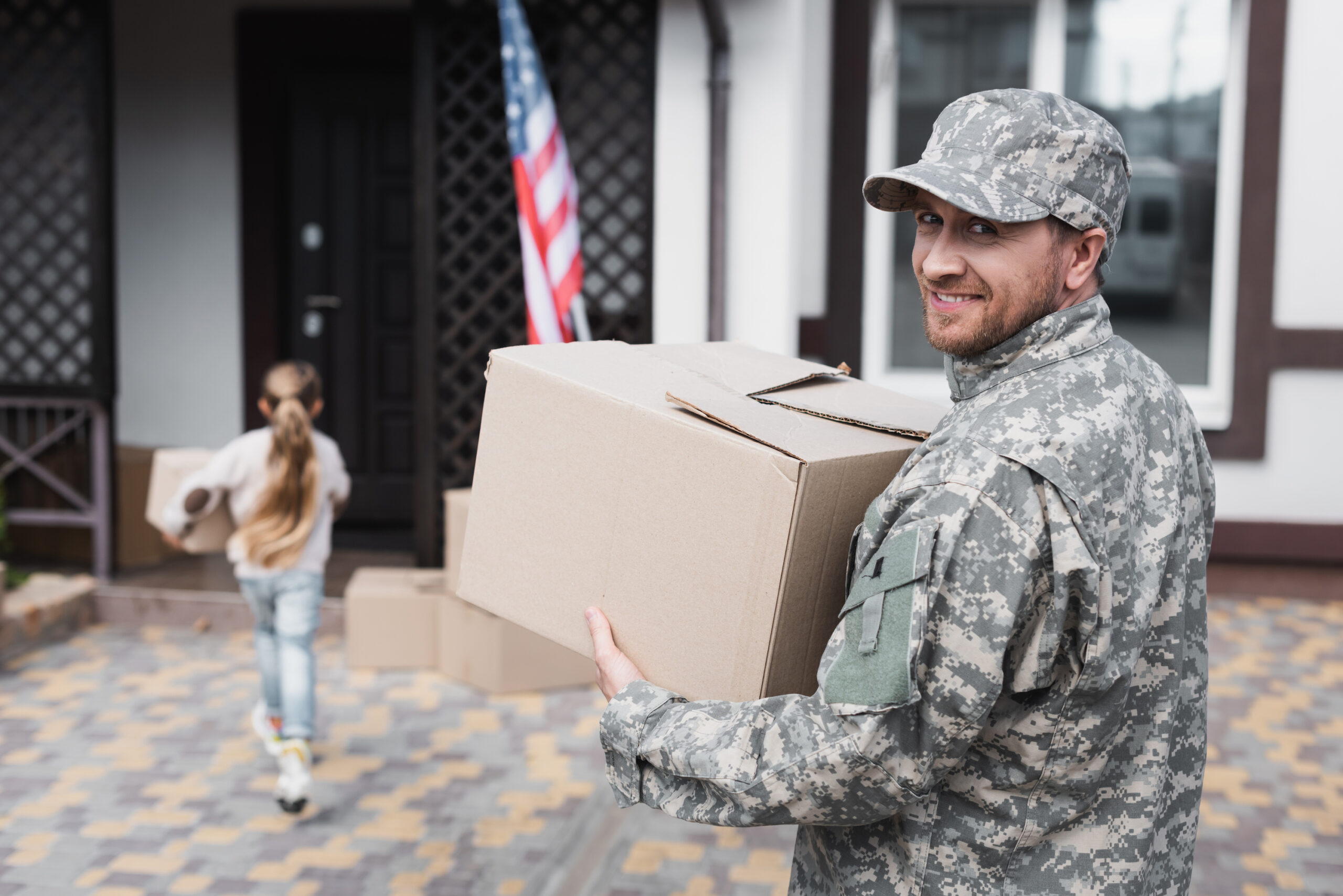 Happy military serviceman holding a cardboard box and looking at camera with girl in the background